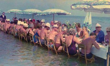 Pranzo sul mare (anzi, nel mare) nell'Italia del Secondo Dopoguerra, qui siamo a Milano Marittima, frazione di Cervia (Ravenna) negli anni Sessanta e lo scatto, di Sante Crepaldi, è tratto da Silvano Collina - Il visionario, libro biografico scritto da Massimo Previato e dedicato appunto a Collina, mitico albergatore "estroso", allora direttore del Grand Hotel Bellevue di Milano Marittima stessa

