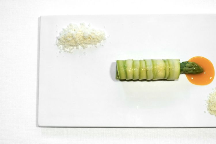 Asparagus with elderberry flower and macadamia nut: the stripe of green asparagus is wrapped around a sorbet of white asparagus. On top, the tip of a green asparagus placed on mandarin juice. Two powders on the side: on top, macadamia, below, a frozen powder of elderberry flower