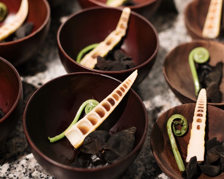Forest Shabu
Una fetta di guanciale di cinghiale cuoce in un brodo caldo e intenso a base di funghi, in particolare orecchie di Giuda che conservano una carnosità incisiva, quasi tendente al croccante; poi il tocco legnoso del bamboo e la felce, così vicina alla freschezza di un asparago selvatico crudo. Il guanciale apporta all’assaggio una sferzata di sapidità, pepata, fresca. Tutto il gusto del selvatico in una ciotola